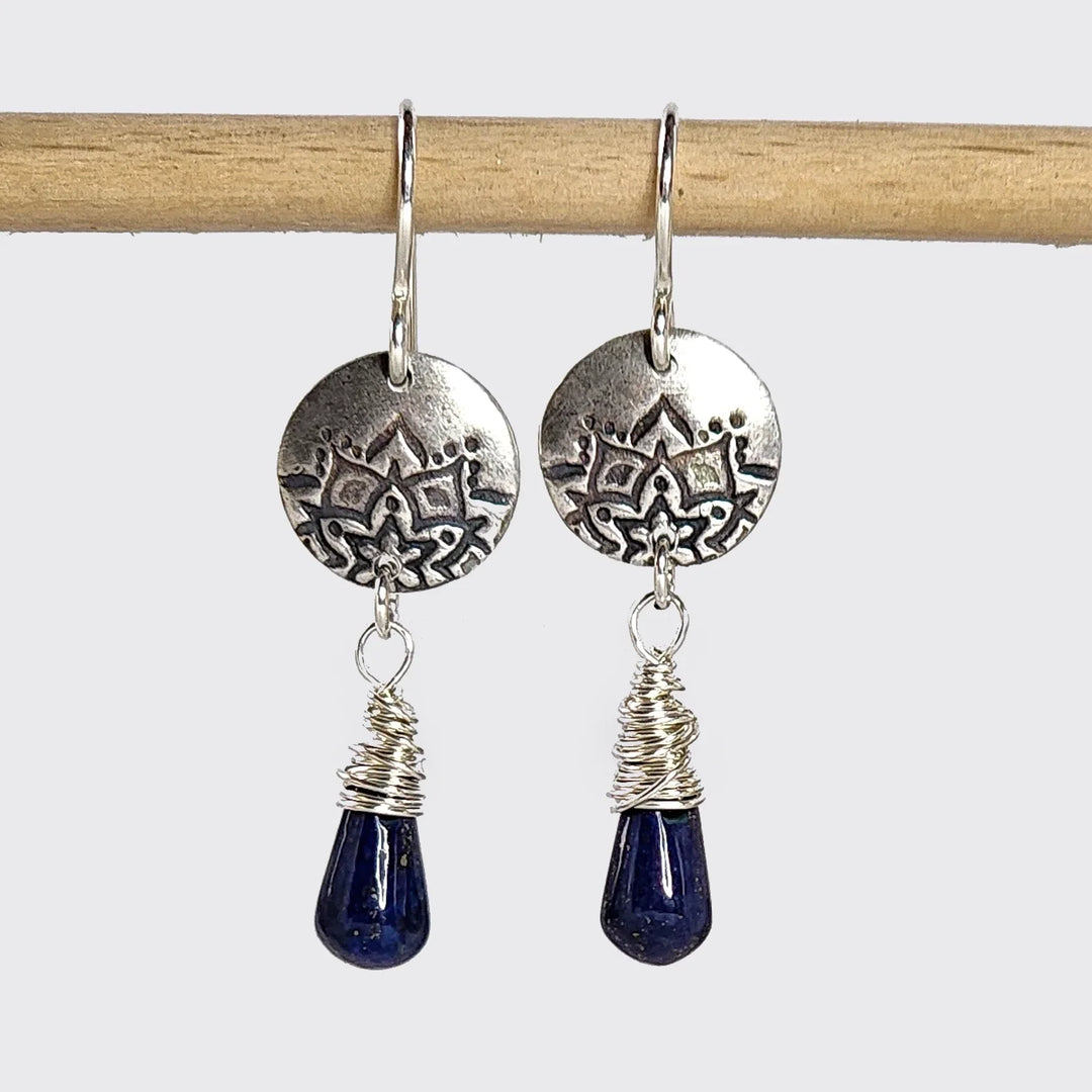 Silver lotus earrings with wire-wrapped lapis stones hanging from a wooden rod.